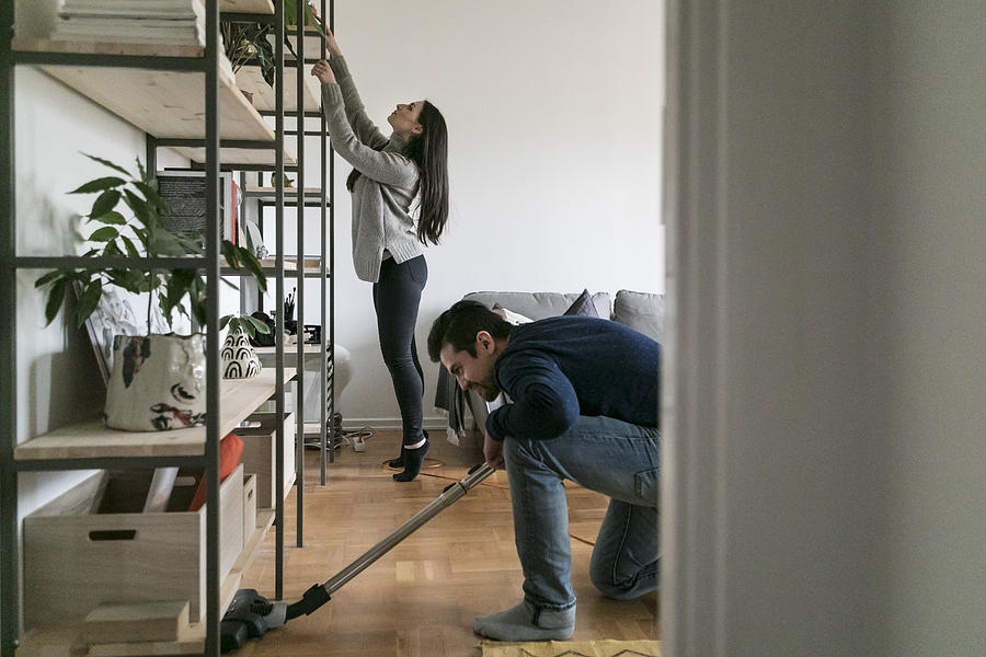 Man vacuuming floor while woman cleaning shelf at home Photograph by Maskot