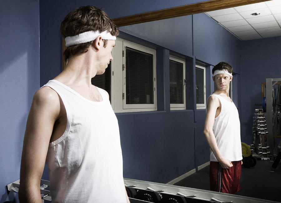 Man wearing sweatband looking at reflection in gym mirror, flexing arm Photograph by Holloway