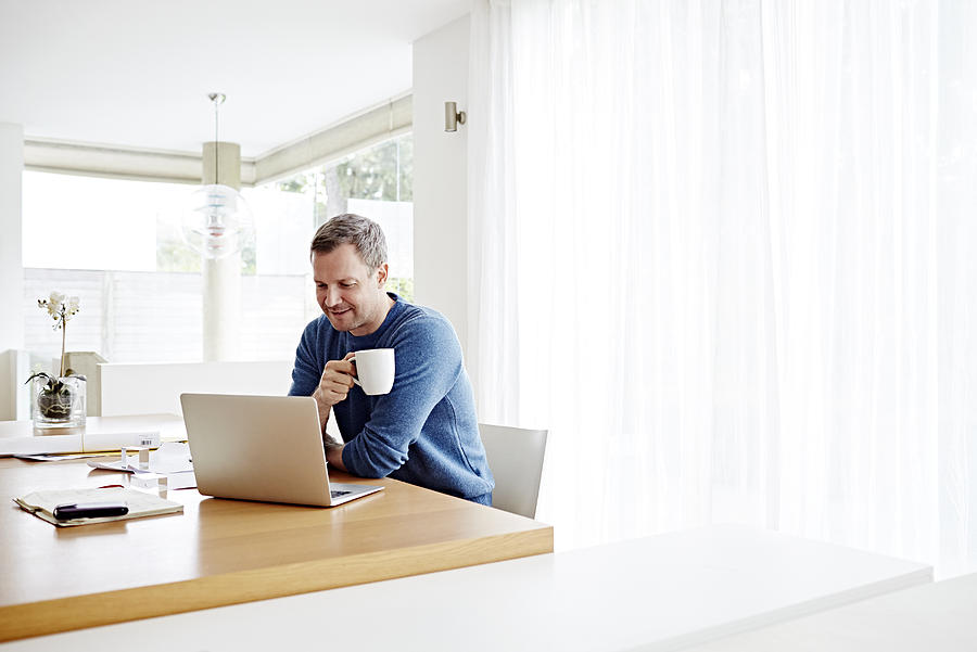 Man working at home using laptop drinking coffee Photograph by Ezra Bailey