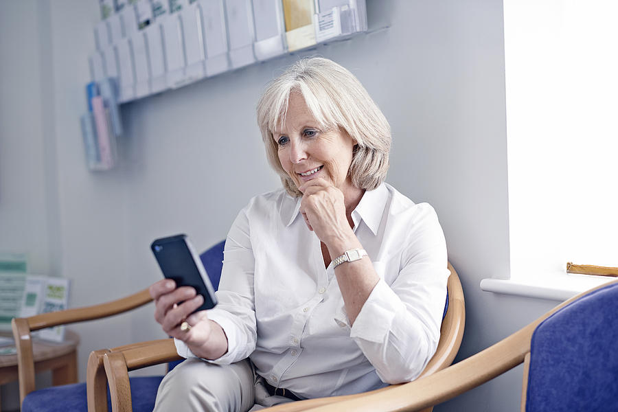 Mature female patient looking at mobile phone in hospital waiting room Photograph by Phil Fisk