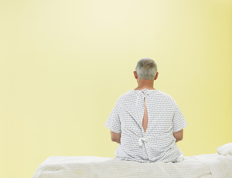 Mature male patient sitting on gurney in hospital gown, rear view Photograph by Siri Stafford