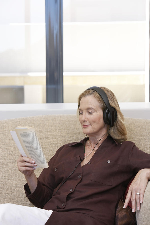 Mature woman sitting on sofa wearing headphones while reading book Photograph by Andrew Olney