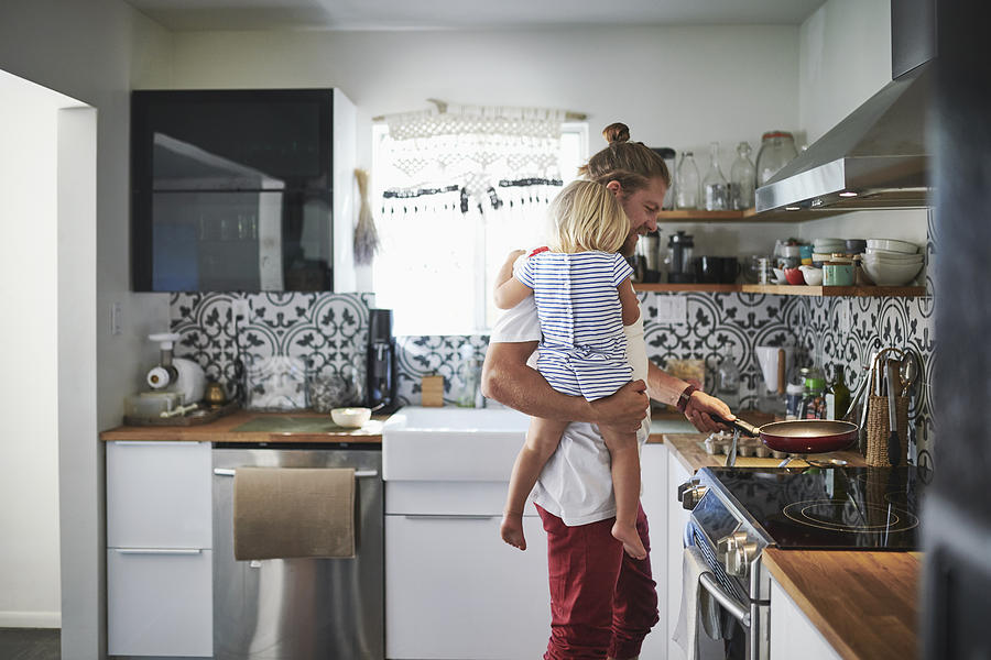 Mid adult father carrying daughter while cooking food in kitchen Photograph by The Good Brigade