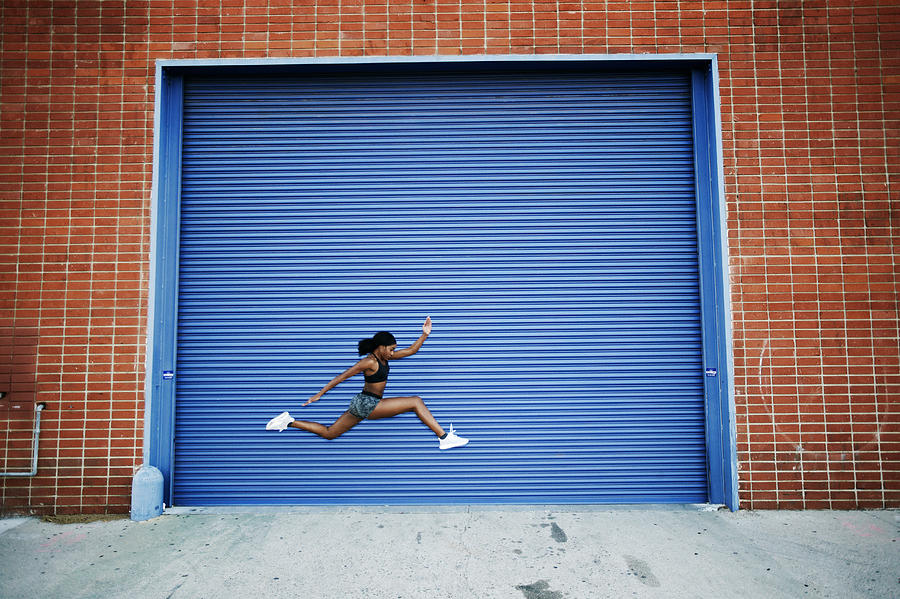 Mixed Race woman running and jumping near blue garage door Photograph by Peter Griffith