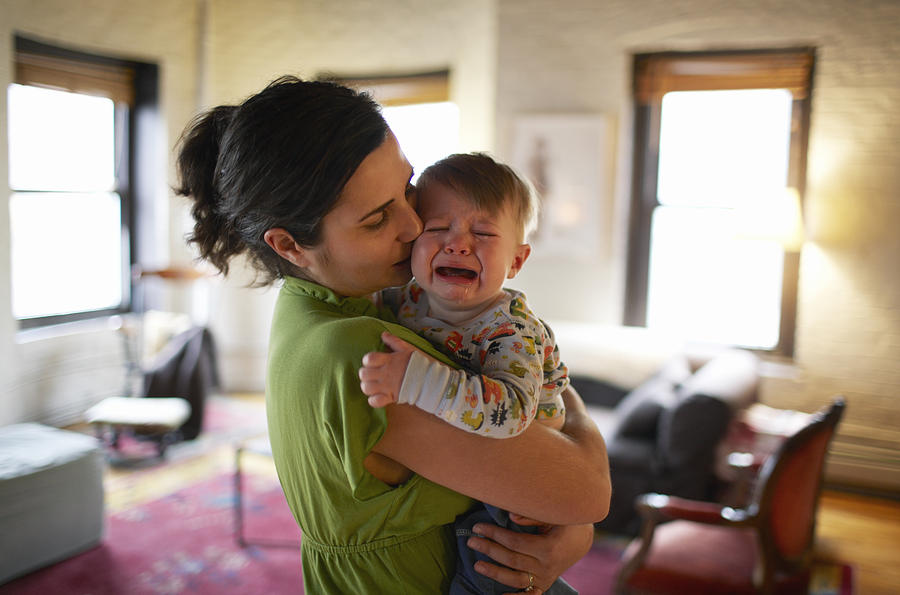 Mother holding and kissing crying baby boy (12-17 months) (focus on foreground) Photograph by Ben Bloom