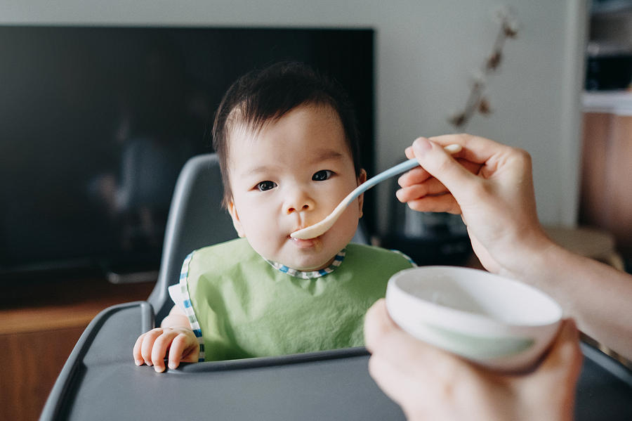 Mothers hand feeding baby girl food on high chair at home Photograph by D3sign