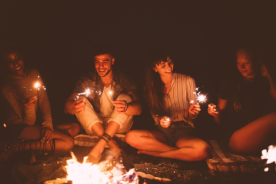 Multi-ethnic hipster friends celebrating with sparklers sitting around beach campfire Photograph by Wundervisuals