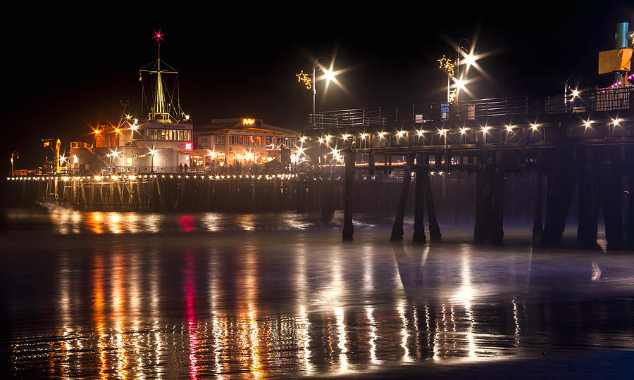 Night On Santa Monica Beach Pier With Bright Colorful Lights Reflecting On The Ocean and Sand Fine A Photograph by Jerry Cowart