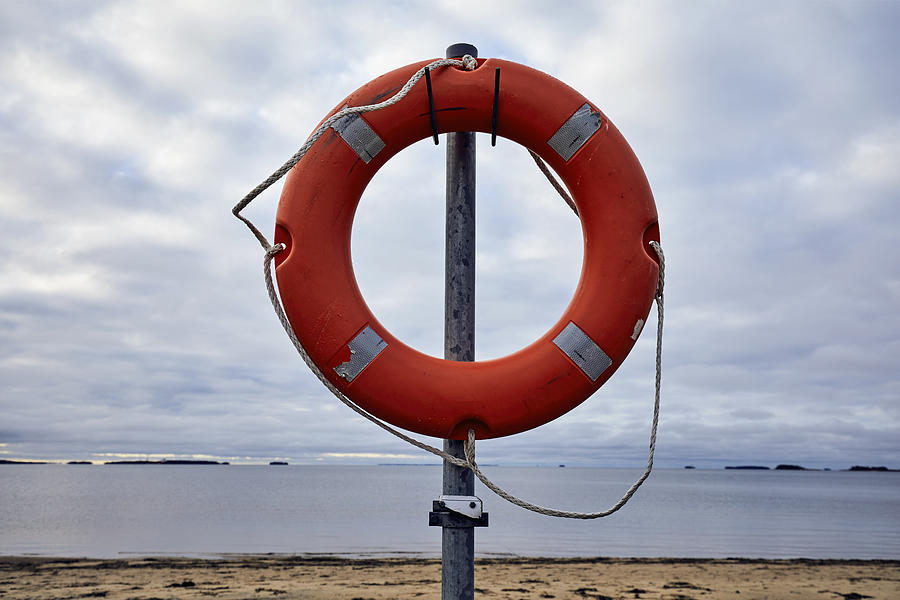 Orange ring buoy with rope on a pole. In the background is the beach, sea and an overcast sky, Oulu, Finland Photograph by Andrew Merry