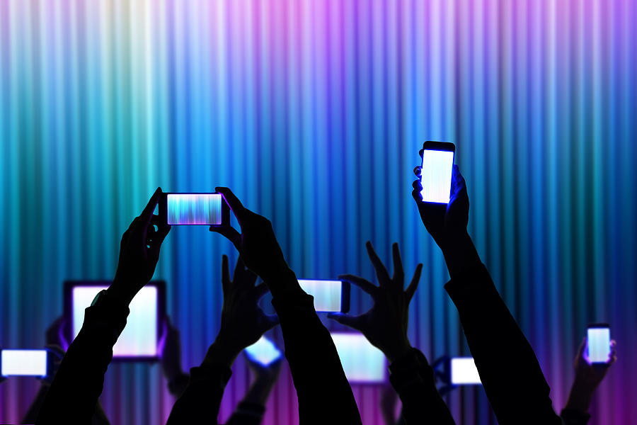 People raise his bright smartphone and tablet device during a night show celebration with dark silhouettes and colorful background. Photograph by Artur Debat