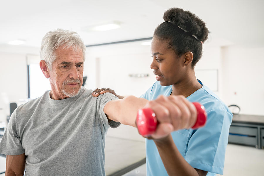 Physiotherapist correcting her senior patient with his shoulder posture as he lifts free weights Photograph by Andresr