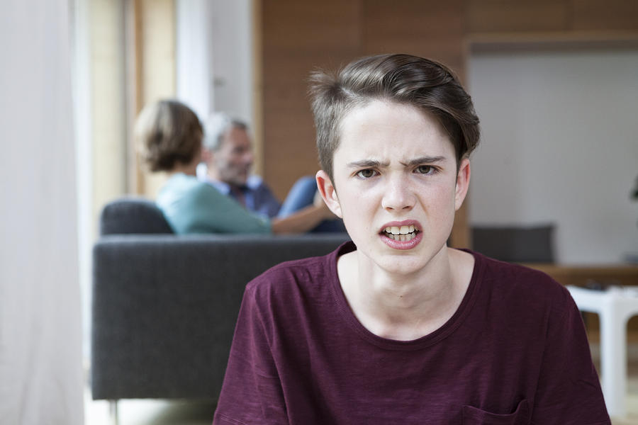 Portrait of angry teenage boy at home with parents in background Photograph by Westend61