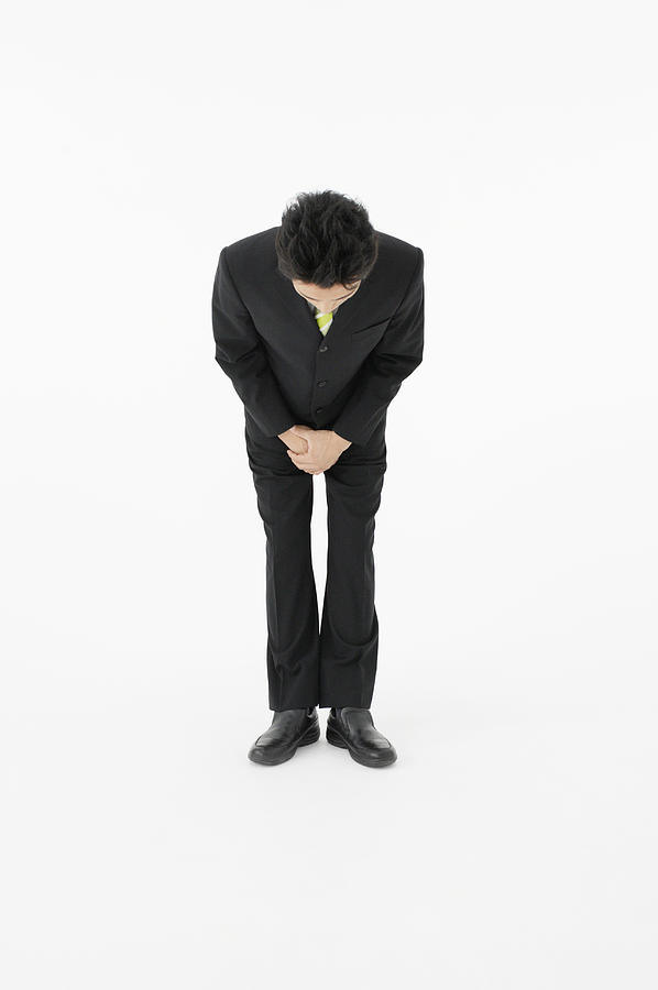 Portrait of businessman bowing with hands clasped, studio shot Photograph by BLOOMimage