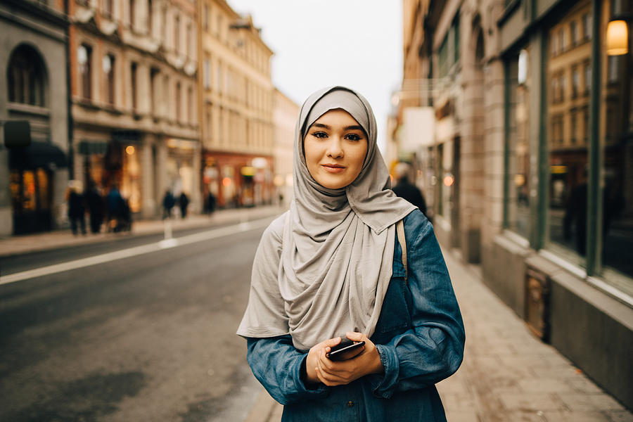 Portrait of confident young woman wearing hijab standing with mobile phone on sidewalk in city Photograph by Maskot