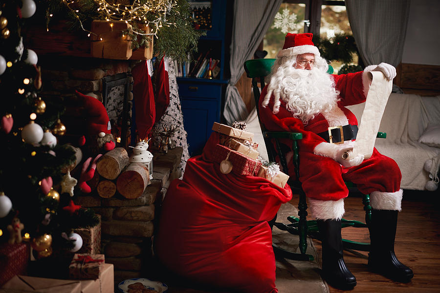 Portrait of Santa Claus, sitting in chair with sack full of presents, looking at Christmas list Photograph by Gpointstudio