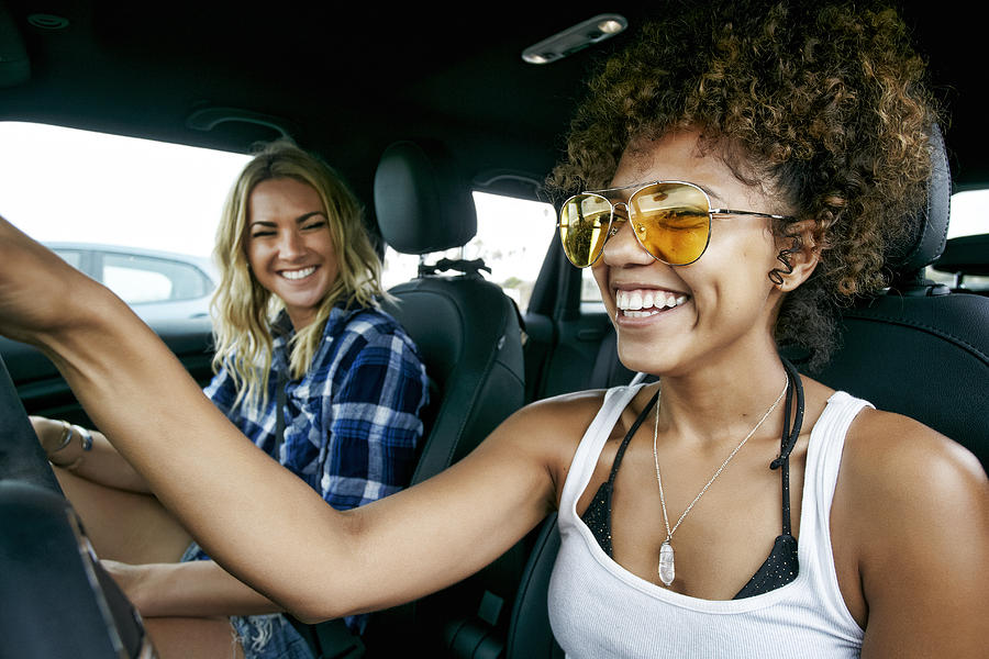 Portrait of two women with long blond and brown curly hair sitting in car, wearing sunglasses, smiling. Photograph by Mint Images