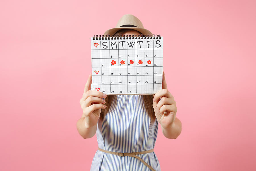 Portrait of woman in blue dress cover face, hiding behind periods calendar for checking menstruation days isolated on trending pink background. Medical, healthcare, gynecological concept. Copy space. Photograph by ViDi Studio