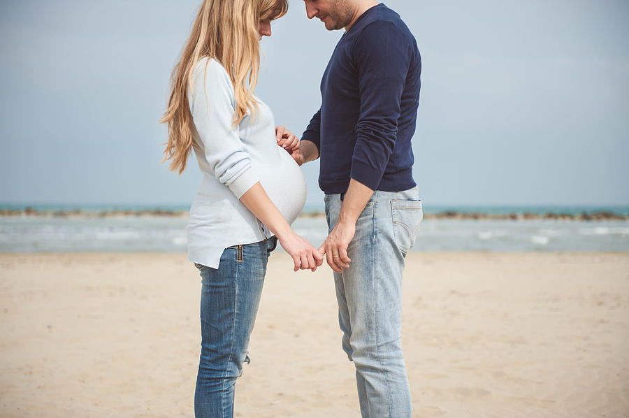 Pregnant couple on the beach. Hands on the belly. Casual clothes. Hand in hand. Photograph by © Samantha Carrirolo