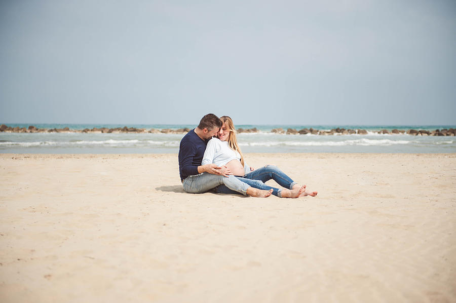 Pregnant couple sitting on the beach. Embrace. Casual clothes. Photograph by © Samantha Carrirolo