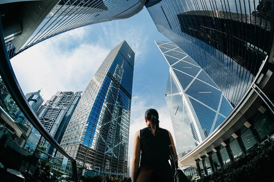 Rear view of professional young businesswoman standing against contemporary financial skyscrapers in downtown financial district and looking up into sky with positive emotion Photograph by D3sign