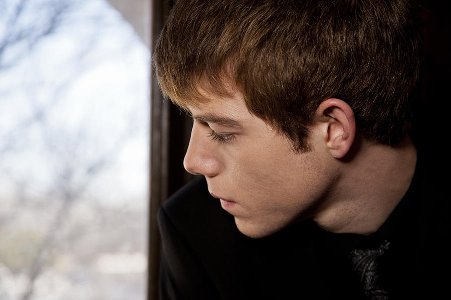 Sad, lonely teenage boy looking out window. Funeral, loss, grief. Photograph by Fstop123