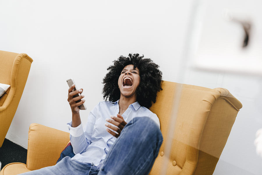 Screaming young woman with cell phone sitting in armchair Photograph by Westend61