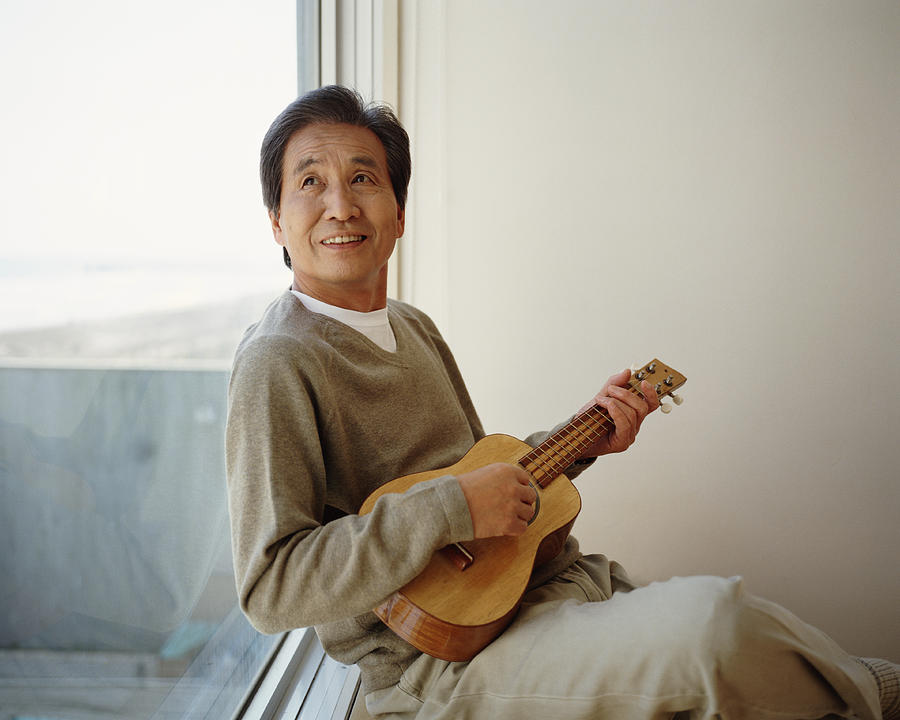 Senior man sitting by window playing ukelele, looking up Photograph by Digital Vision