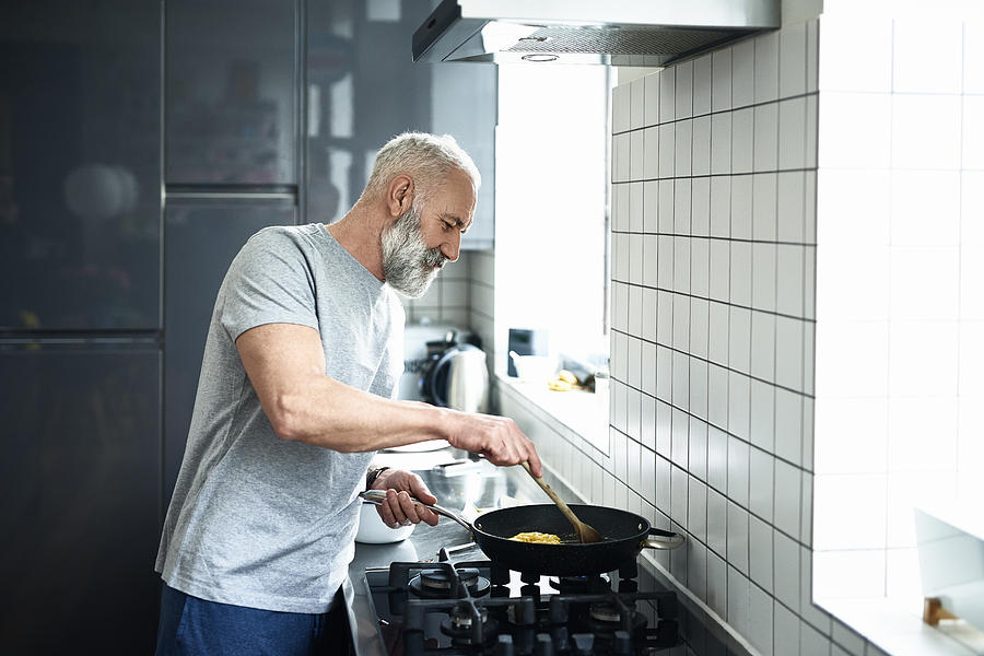 Senior man with grey beard using frying pan in modern kitchen Photograph by 10000 Hours