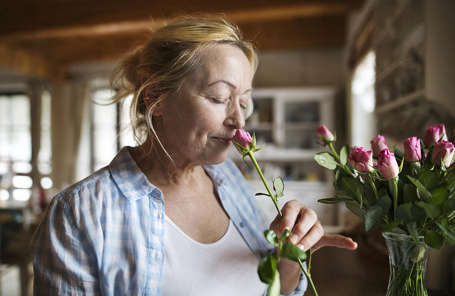 Senior woman in her kitchen smelling fragrance of pink rose Photograph by GrapeImages