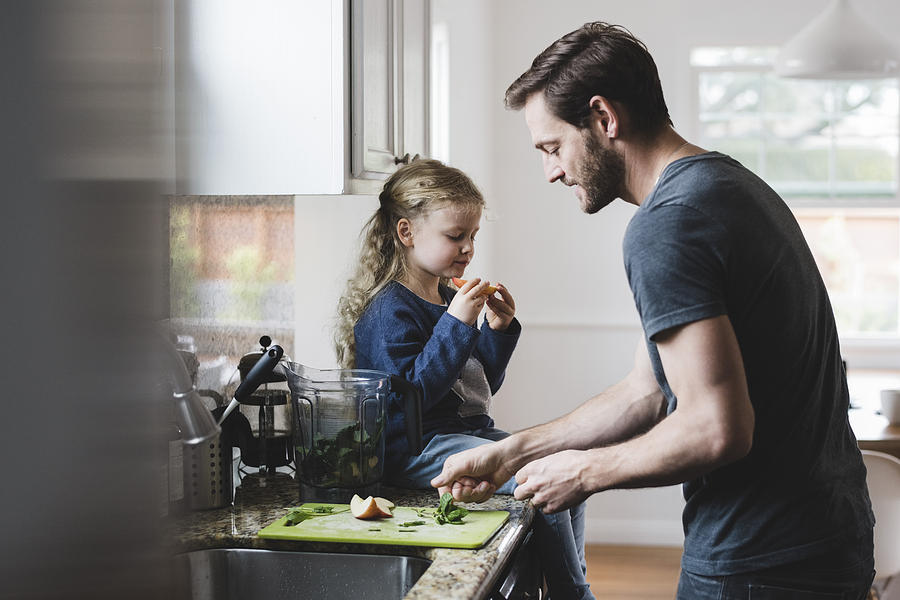 Side view of father cooking food while daughter having apple in kitchen Photograph by Maskot