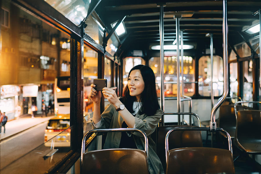 Smiling female tourist taking pictures of night scene with smartphone on a tram ride in Hong Kong Photograph by D3sign