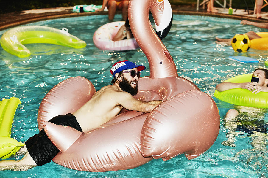 Smiling man hanging out on inflatable pool toy during party with friends on summer evening Photograph by Thomas Barwick