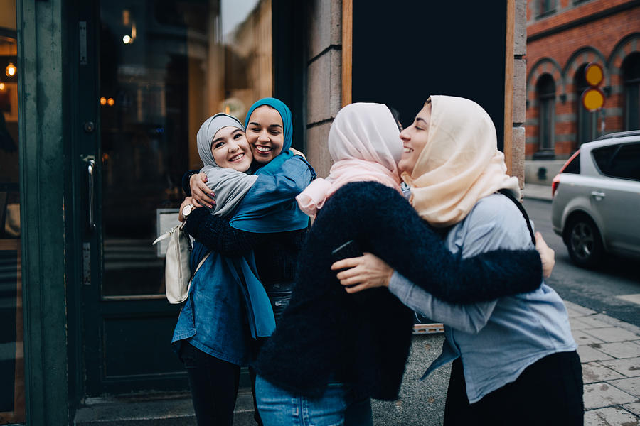 Smiling multi-ethnic female friends greeting on sidewalk in city Photograph by Maskot