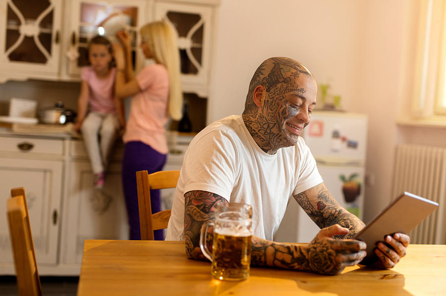 Smiling tattooed man using touchpad in the kitchen. Photograph by Skynesher