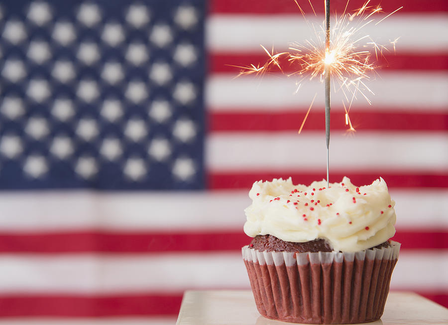 Studio shot of sparkler atop cupcake, american flag in background Photograph by Jamie Grill