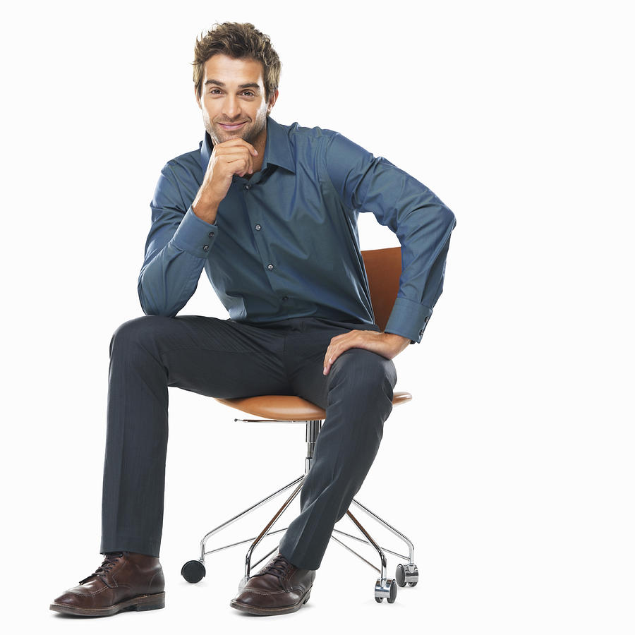 Studio shot of young business man sitting on chair with hand on chin and smiling Photograph by Momentimages
