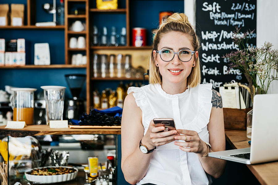 Stylish Young Woman Smiling Using Smartphone In Cafen Photograph by Tom Werner