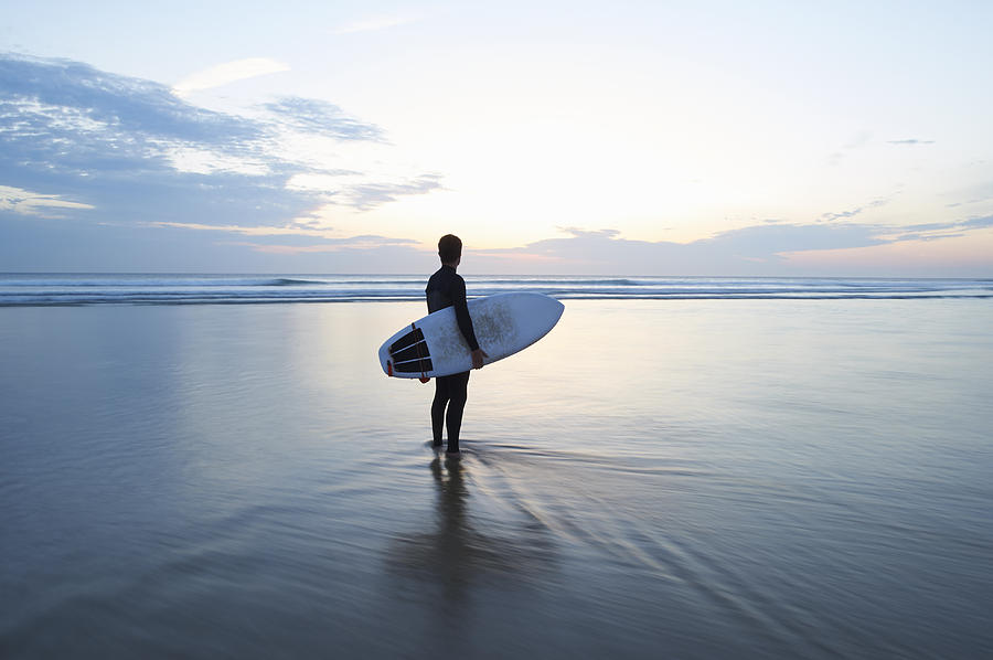 Surfer with surfboard looking out to sea at dusk. Photograph by Dougal Waters