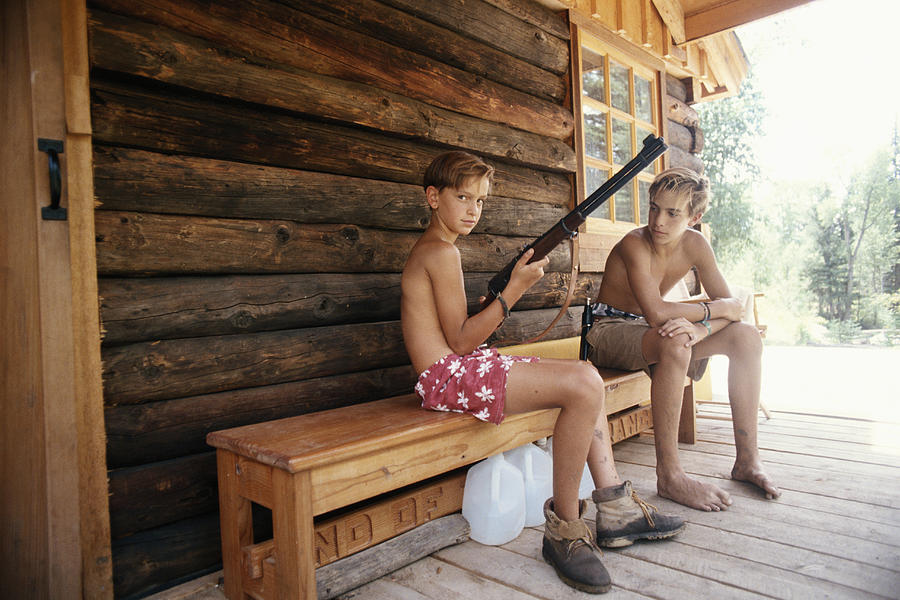 Teenage boy (13-15) and boy (10-13) sitting on bench in porch, boy holding rifle Photograph by David De Lossy