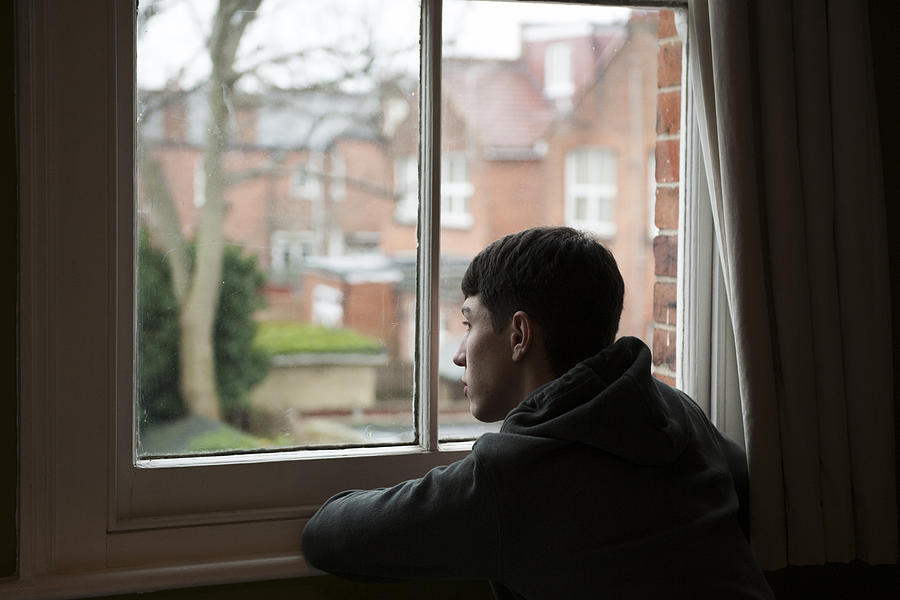 Teenage boy looking out of bedroom window Photograph by Richard Bailey