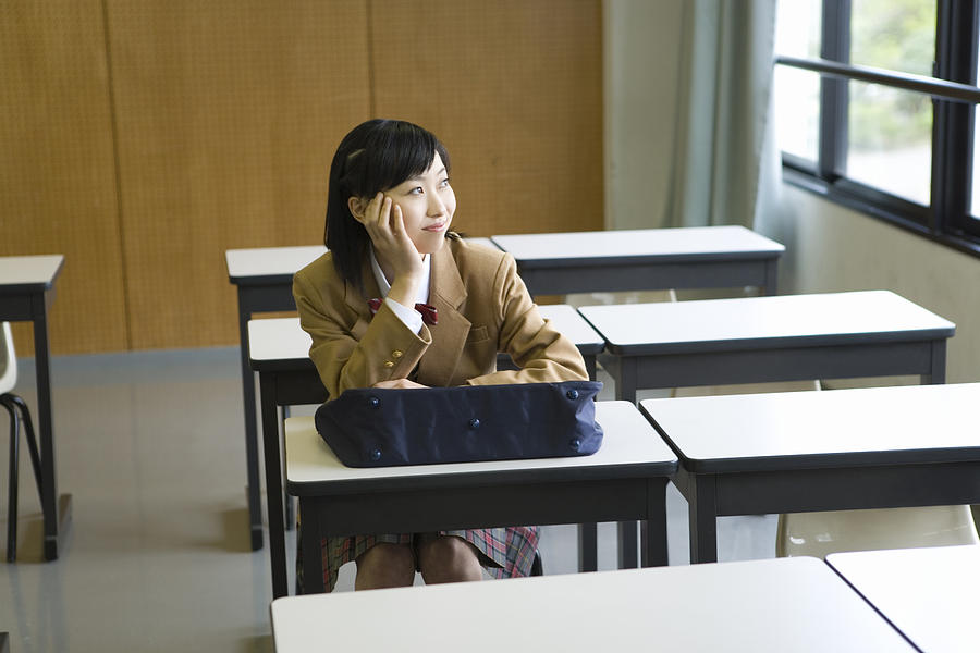 Teenage girl smiling and sitting at desk in classroom, resting chin in hand Photograph by Daj