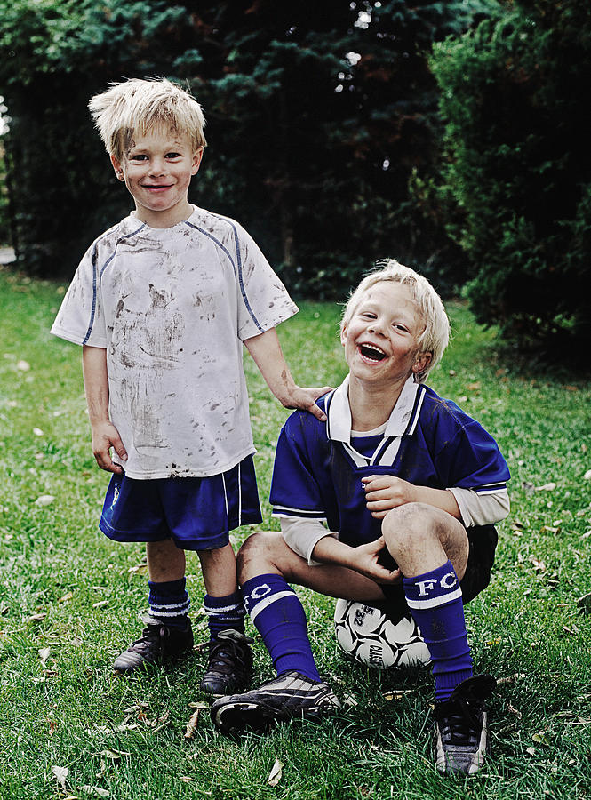 Twin boys (6-8) wearing football kits, one sitting on ball, portrait Photograph by David Trood