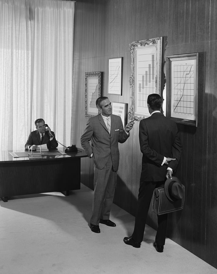 Two businessman discussing at bar chart while another man using telephone in background Photograph by Tom Kelley Archive