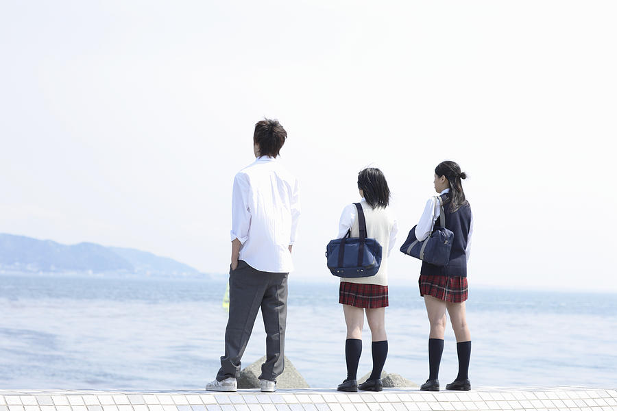 Two female and one male high school students (17-18) looking at sea, rear view Photograph by Sot
