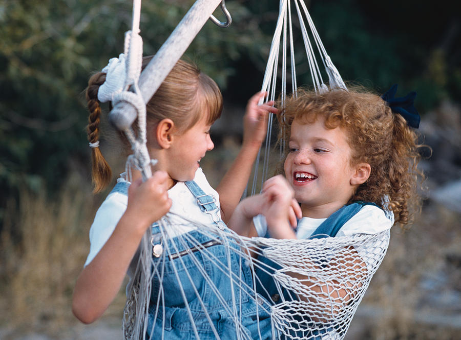Two Little Girls Wearing Denim Overalls And White Shirts Are Playing In A Hammock While Laughing And Smiling At Each Other Photograph by Photodisc