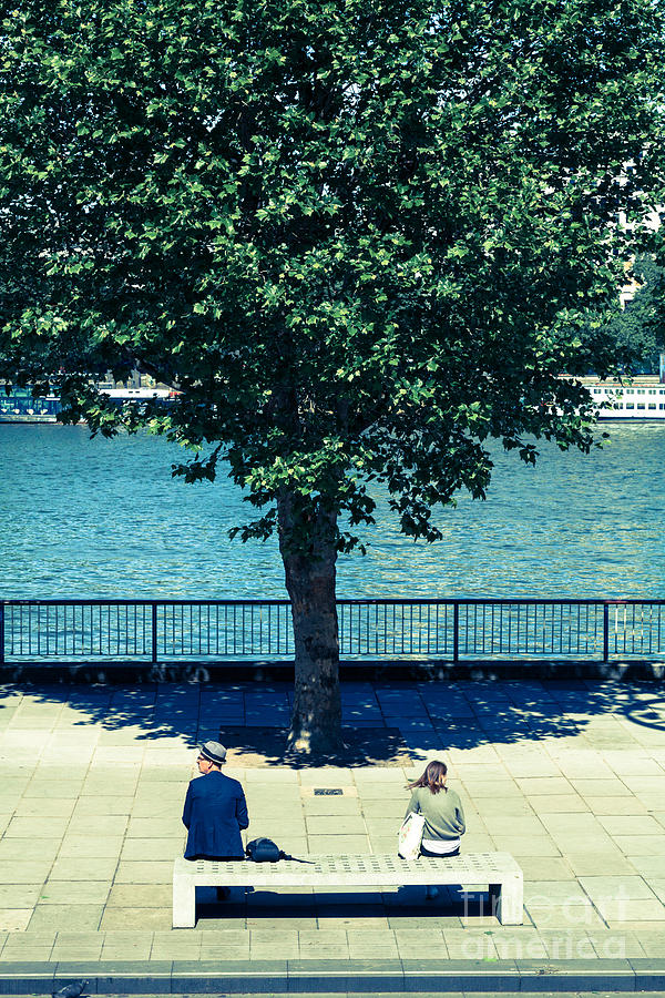 Two people sitting on concrete bench by river with tree. Photograph by Peter Noyce