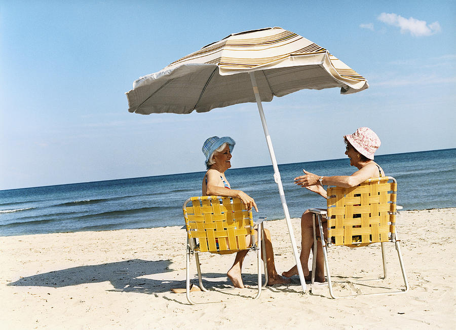 Two Senior Women Sit on Deckchairs Under a Parasol on the Beach, Talking Photograph by Digital Vision.