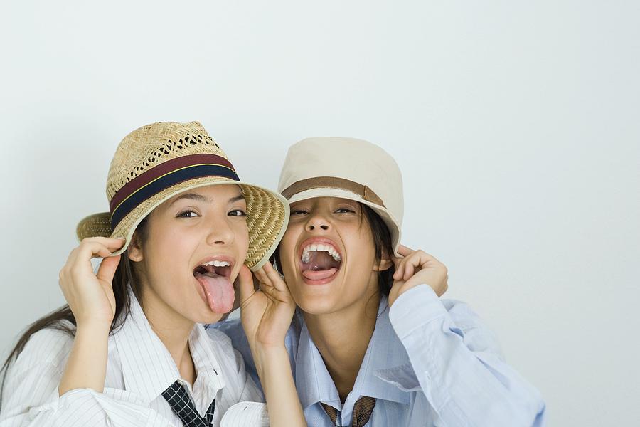 Two young friends wearing hats, sticking out tongues, looking at camera, portrait Photograph by PhotoAlto/Laurence Mouton