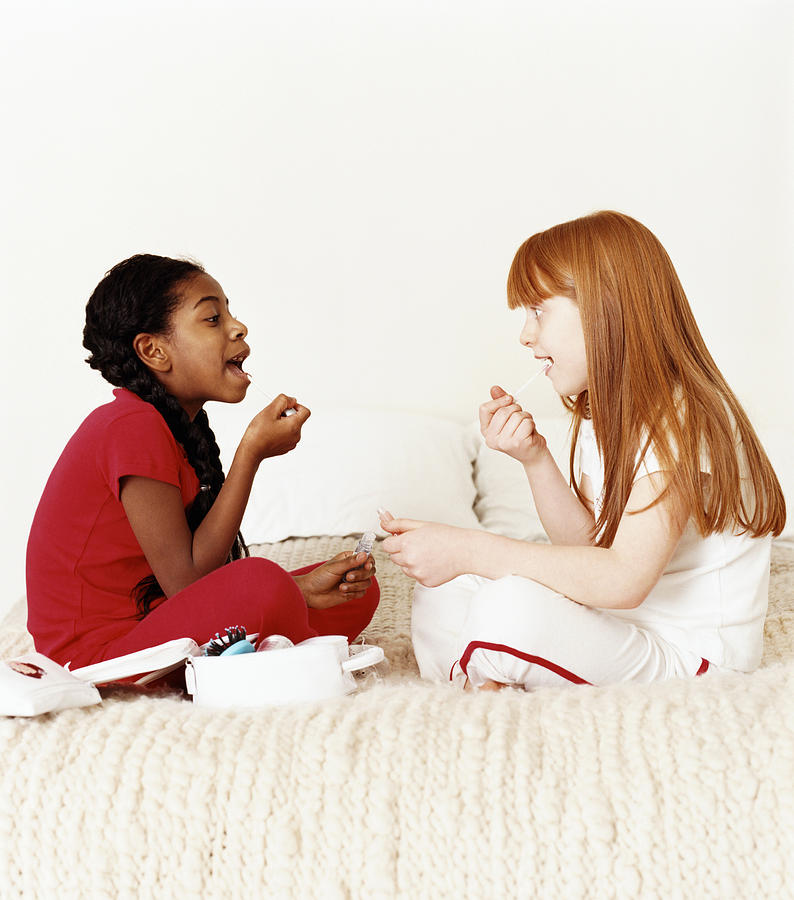 Two Young Girls Sitting Face to Face on a Bed Applying Lip Gloss Photograph by Lottie Davies