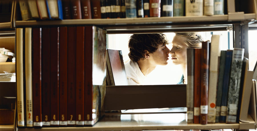 Two Young Students Kissing in a Library, Viewed Through a Gap Between Books on a Shelf Photograph by Digital Vision.